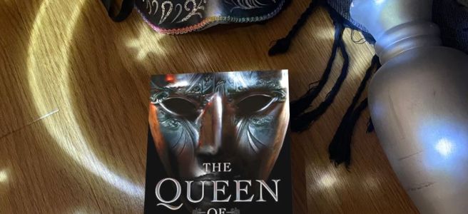 The Queen of Days book cover on a wooden backdrop with a silver column on the right, a mask on the top, and stars and a crescent moon lit up in the backdrop