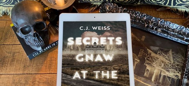 Secrets Gnaw at the Flesh book cover on an iPad above a framed picture of a haunted house, a wooden backdrop, wiht a black skull and lit candle