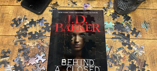 Behind a Closed Door book cover on a wooden backdrop surrounded by loose puzzle pieces, with a black skull and a smartphone