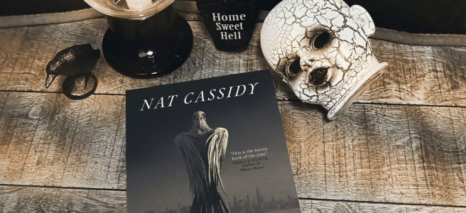 Nestlings book cover on a wooden backdrop, with a little vase that says "home sweet hell", a crow figurine, a creepy baby doll head, and a lit candle
